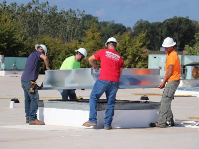 Four Buffington Brothers commercial HVAC technicians in hard hats, one wearing a Buffington Brothers red company shirt, setting curb on a flat commercial rooftop.